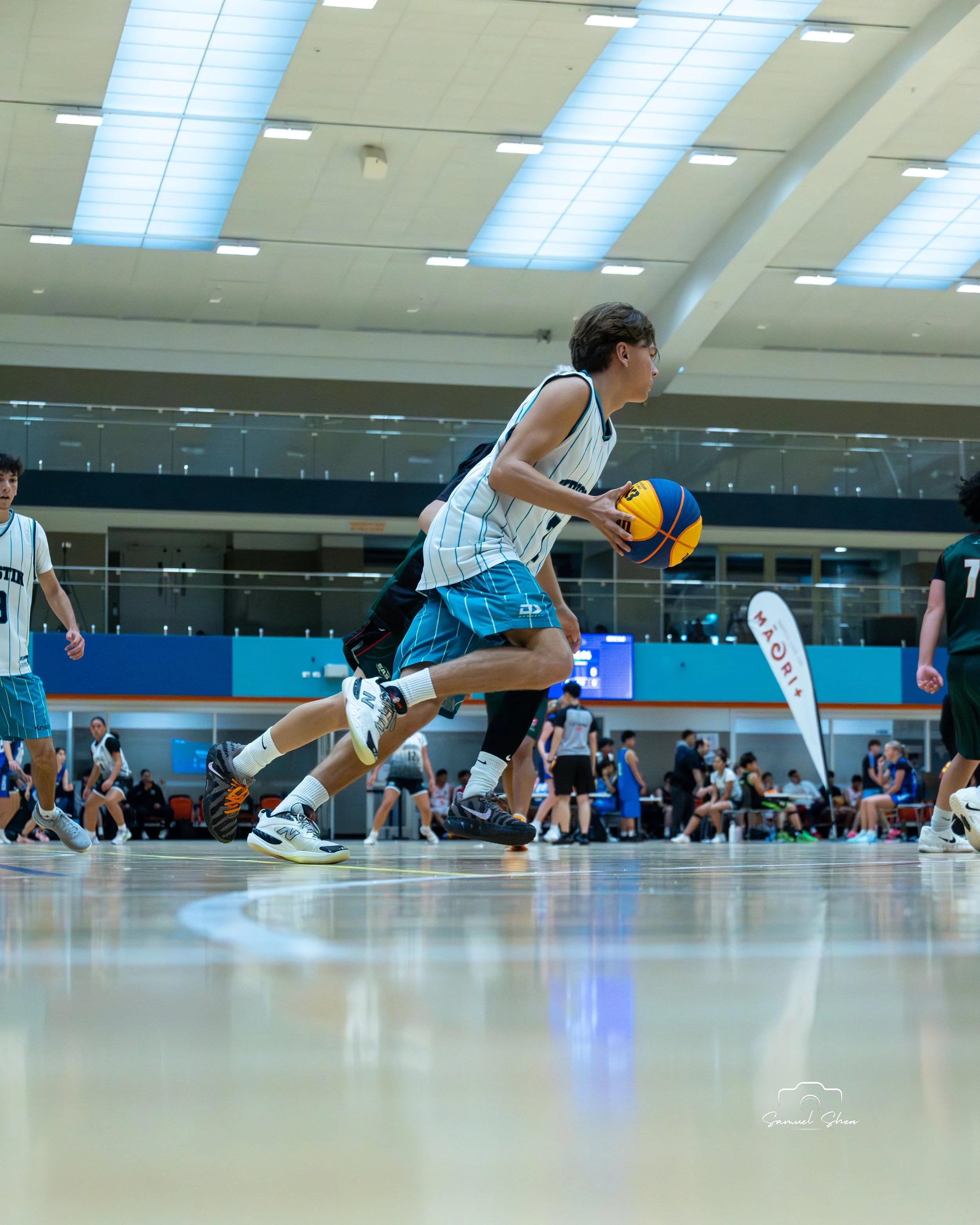A basketball player wearing a white and teal jersey with blue shorts dribbling a blue and yellow basketball on an indoor court. There are other players and people in the background, and the ceiling has bright lights and large skylights.