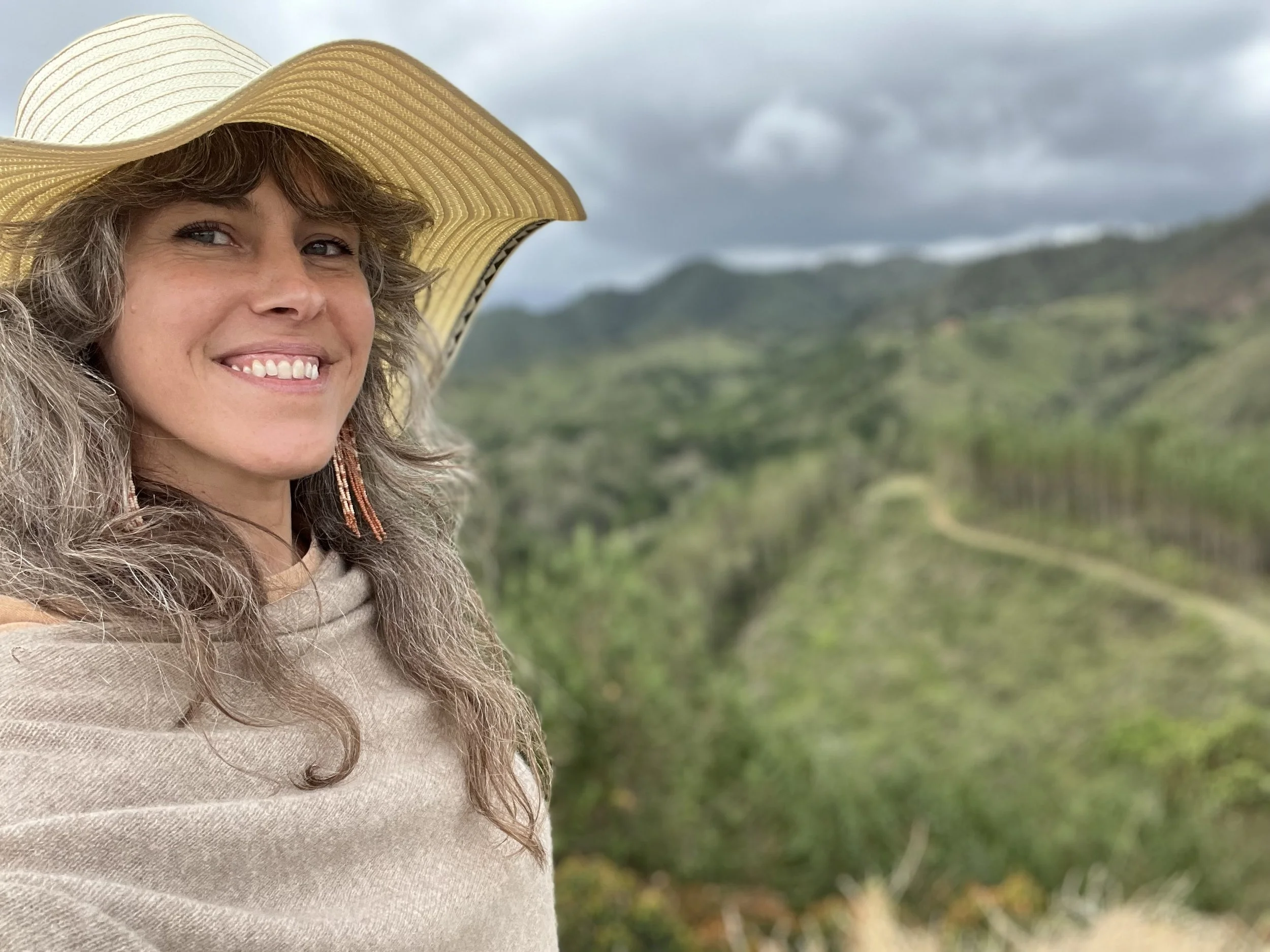 Nova Campbell smiling outdoors wearing a wide brim straw hat with green mountainous landscape behind her