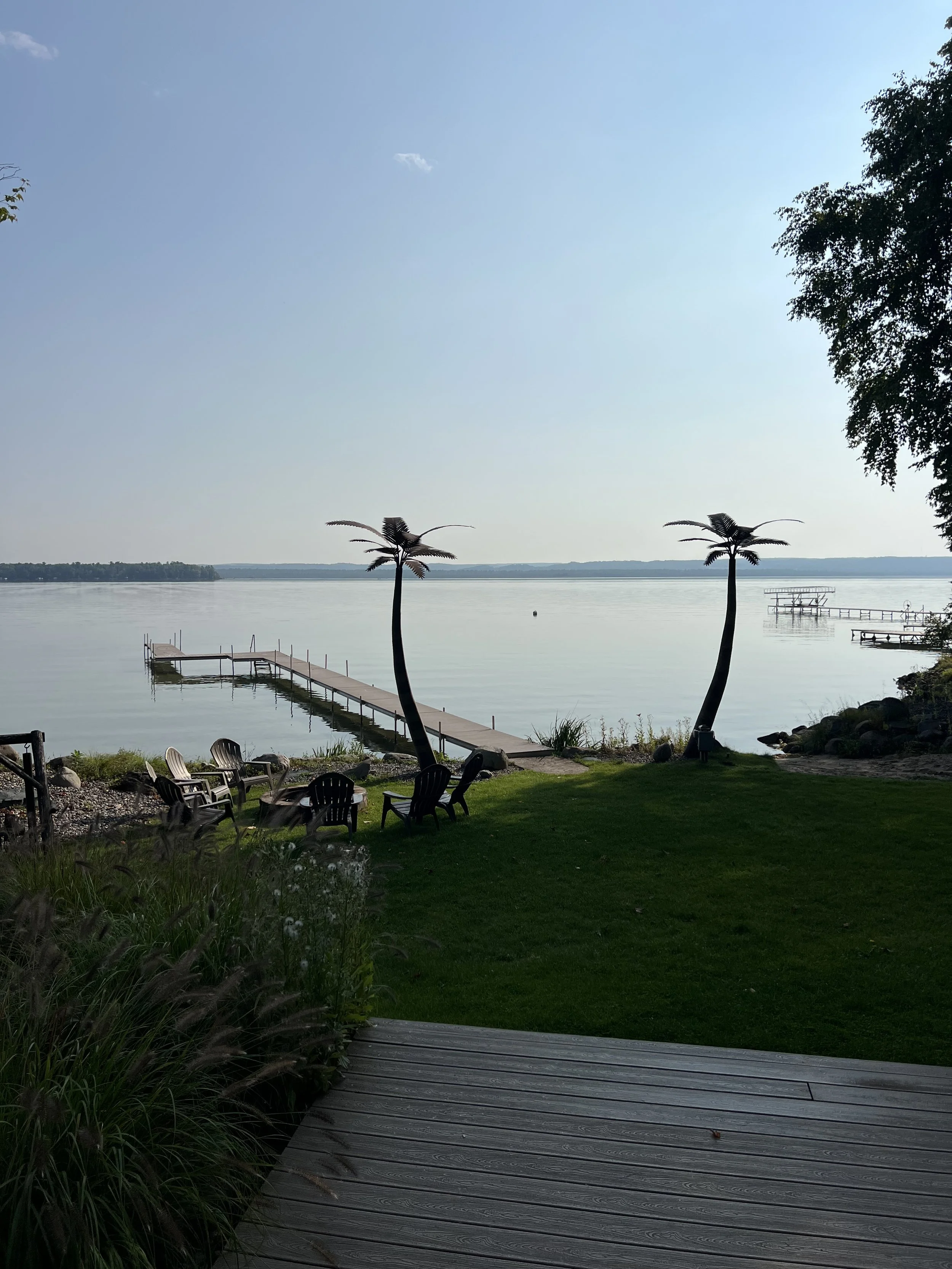A lakeside scene with a wooden dock extending into the water, surrounded by green grass, plants, and trees with two small, decorative palm trees nearby. There are several chairs on the grass, and a floating dock is visible on the water in the background.
