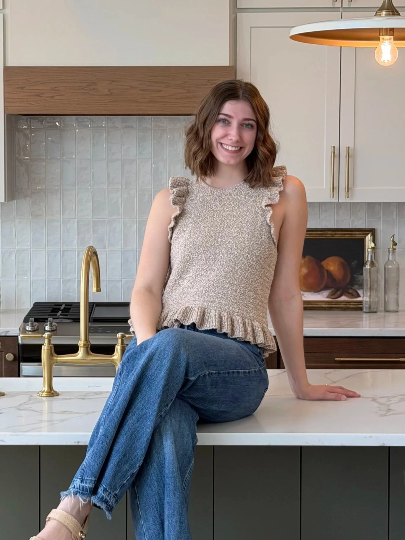 A woman sitting on a kitchen counter, smiling, wearing a ruffled sleeveless beige top and blue jeans. Behind her is a modern kitchen with white cabinets, a gold faucet, a black stove, and decorative glass bottles. A framed painting of peaches is on the wall, and a modern pendant light hangs above.