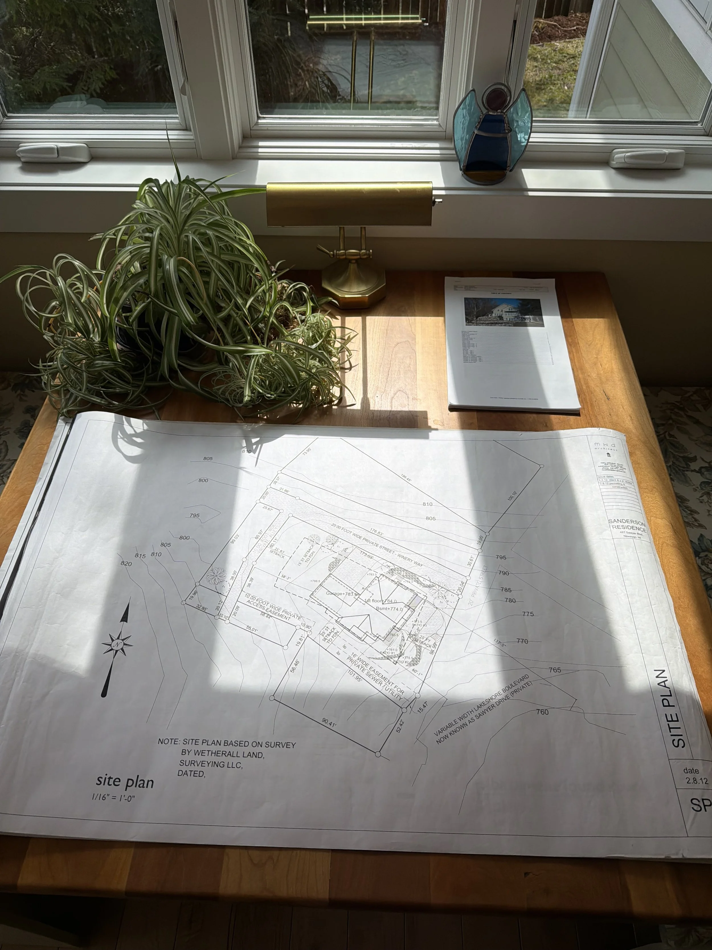 A detailed site plan drawing of a property at a desk with sunlight coming through a window, a potted spider plant, a brass desk lamp, a small decorative stained glass butterfly ornament, and a folded stack of papers.