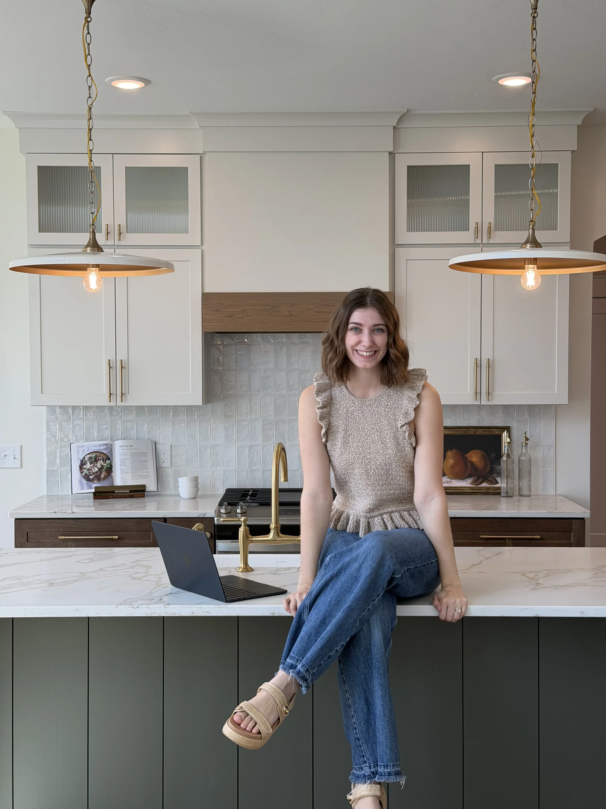 A young woman with brown hair, wearing a beige sleeveless top and blue jeans, sitting on a white kitchen counter with a laptop open in front of her. The kitchen has white upper cabinets with glass doors, brass handles, and a marble countertop. Two hanging pendant lights with brass fixtures and exposed light bulbs are above her.