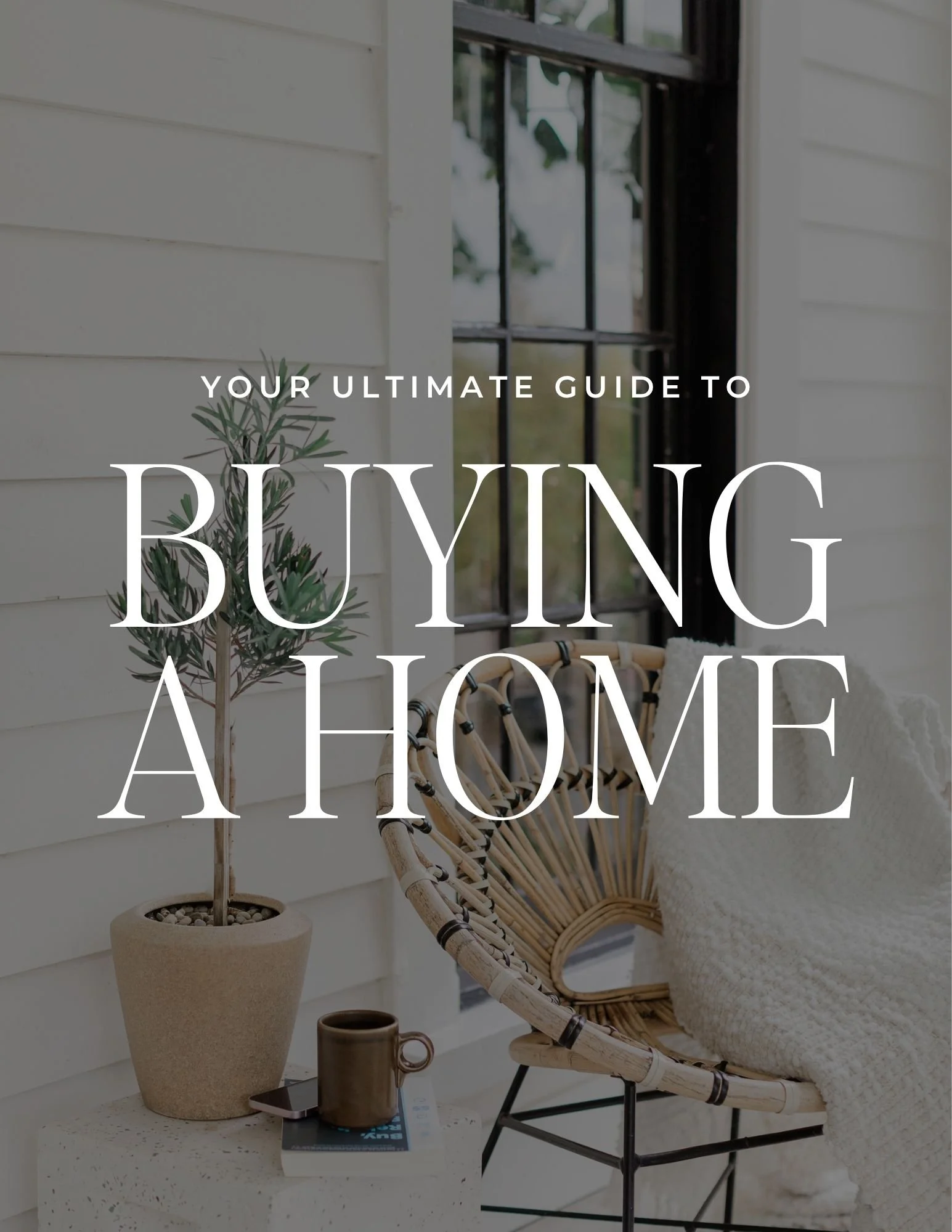A cozy porch with a small potted olive tree, a tablet, a coffee mug, and a book on a speckled white table. There is a rattan chair with a cream blanket and black metal accents, set against white siding and a black-framed window.