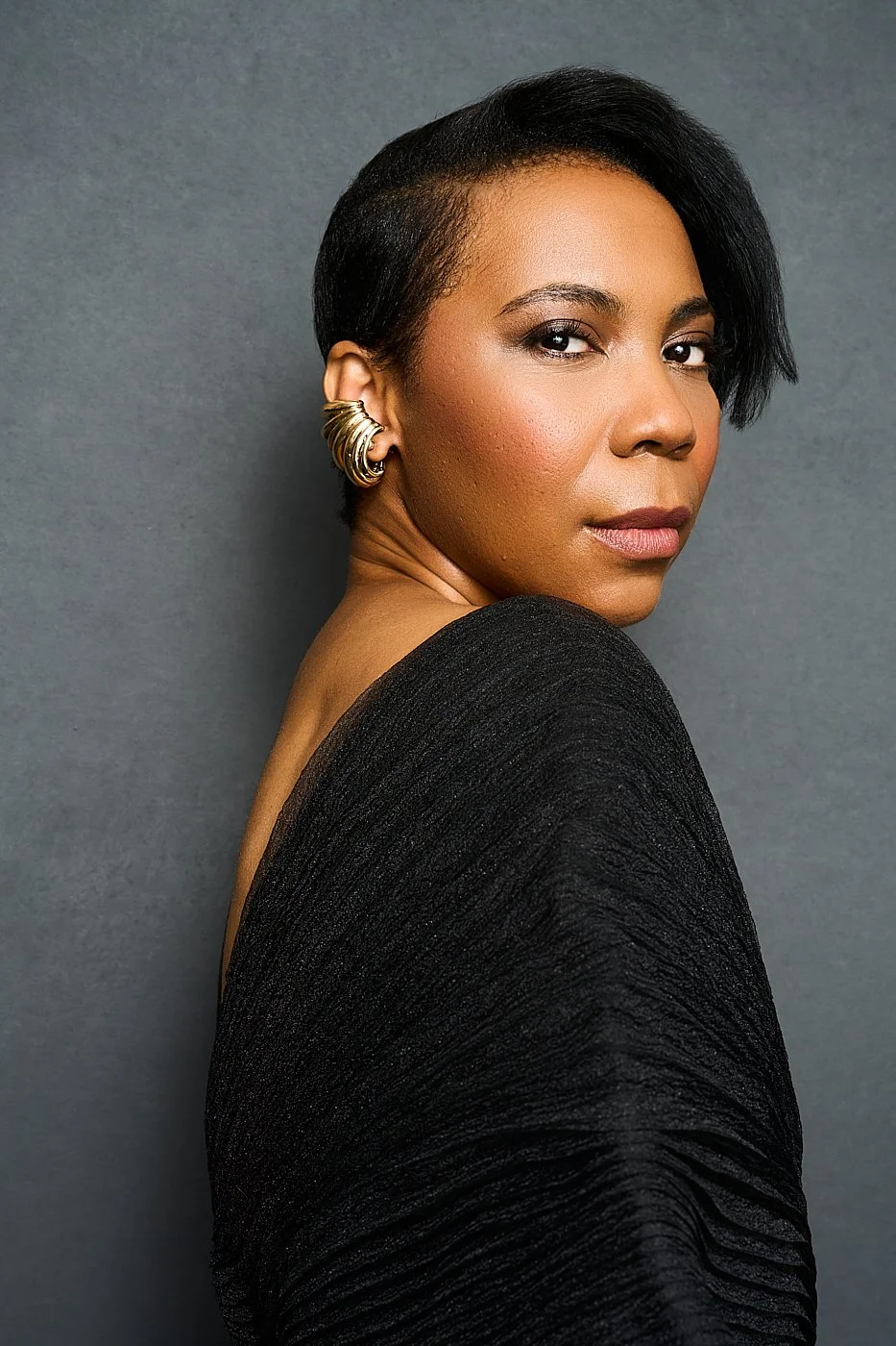 A confident woman with short, styled black hair, wearing large gold hoop earrings and a black textured off-shoulder top, looking over her shoulder at the camera against a neutral gray background.