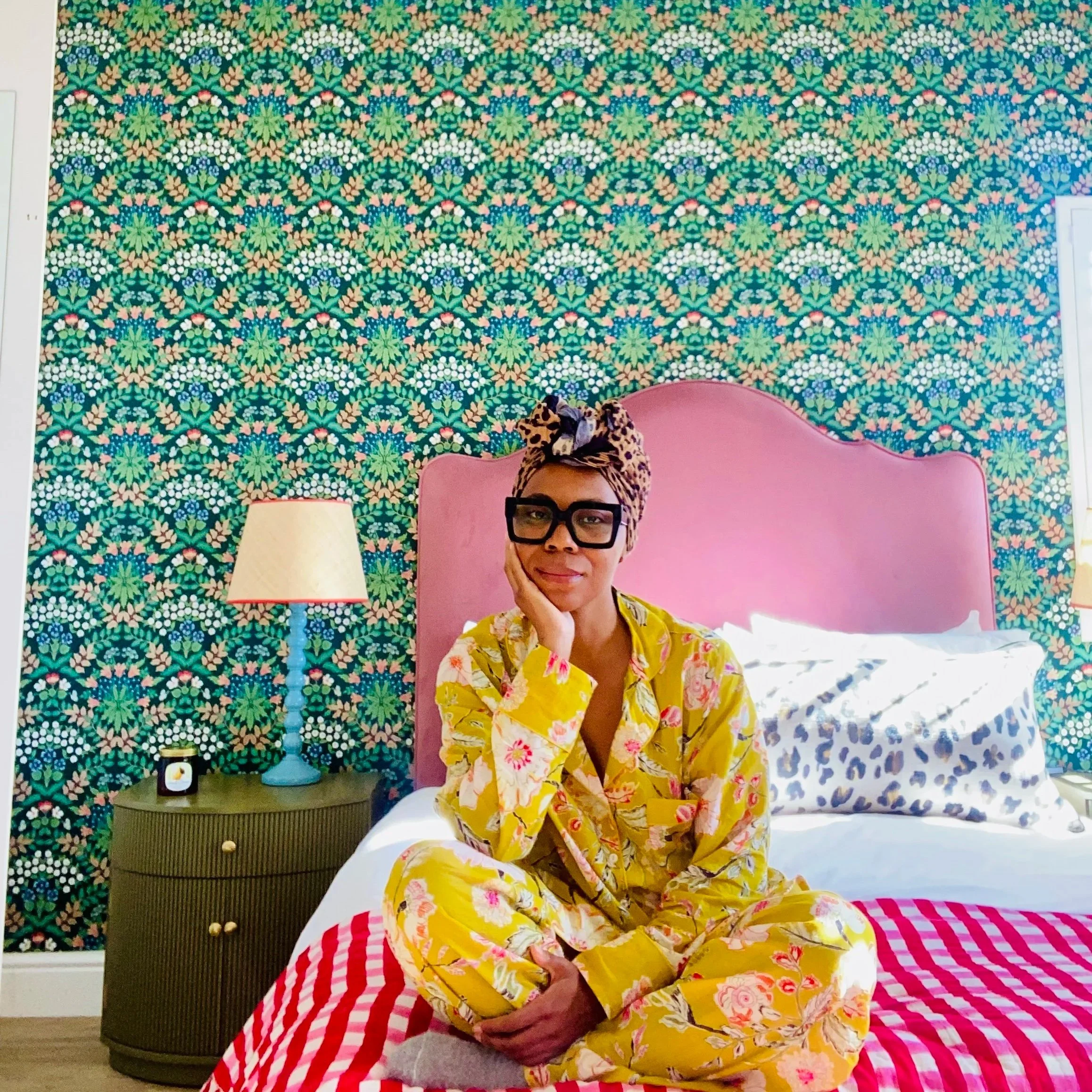 A woman sitting on a bed with a pink headboard, wearing yellow floral pajamas and a leopard print headscarf. She has large glasses and is resting her chin on her hand. The background features a colorful floral-patterned wall, a nightstand with a lamp, a small clock, and a cheetah print pillow on the bed.