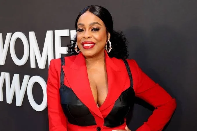 A woman with long black curly hair, dressed in a red blazer with black accents, smiling and posing in front of a backdrop with white text.