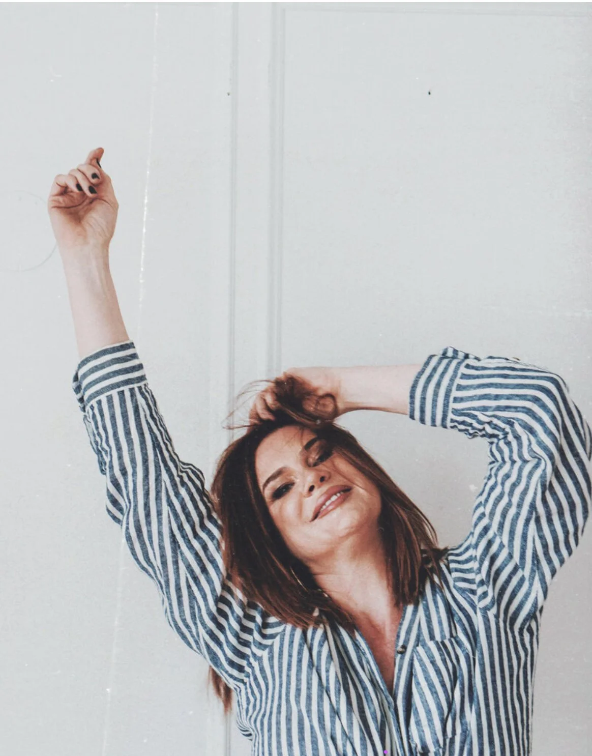 A woman with shoulder-length brown hair, wearing a striped blue and white shirt, smiling with her eyes closed, with one hand on her head and the other arm raised.