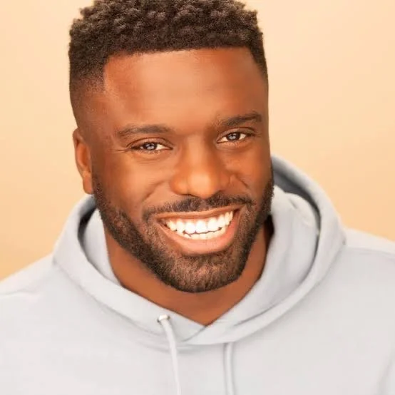 Close-up portrait of a smiling man with dark skin, short curly hair, beard, wearing a light gray hoodie, against a beige background.
