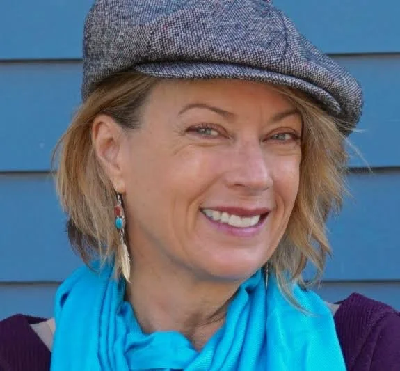 A woman with blonde hair wearing a gray flat cap, turquoise earrings, and a bright blue scarf, smiling in front of a blue wall.