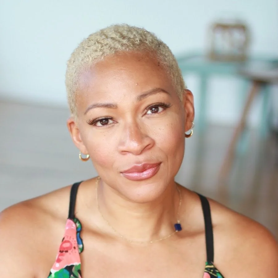 A woman with short curly platinum blonde hair, wearing a floral top with black straps, gold hoop earrings, and a delicate necklace with a dark blue pendant, sitting indoors with a blurred background.