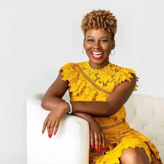 A woman with short, curly blonde hair sitting on a white couch, wearing a bright yellow lace dress, smiling with red lipstick, hoop earrings, and colored nails.