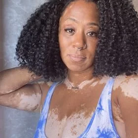 A woman with curly black hair wearing a blue top, taking a selfie in front of a mirror.