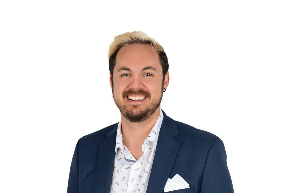 A man with a beard, blonde and dark hair, wearing a blue blazer and a patterned white shirt, smiling against a white background.