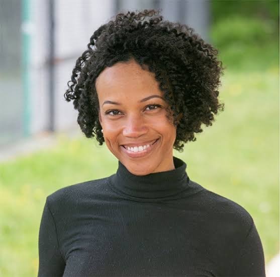 A woman with curly black hair, wearing a black turtleneck, smiling outdoors with green background.