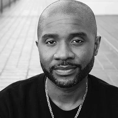 Close-up portrait of a man with a shaved head, beard, and mustache, wearing a black shirt and a chain necklace, smiling softly.