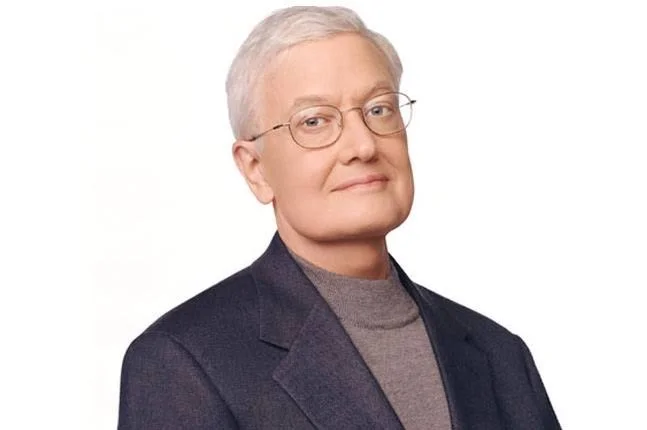 Older woman with short gray hair, glasses, wearing a dark blazer and gray turtleneck, looking confidently at the camera against a white background.