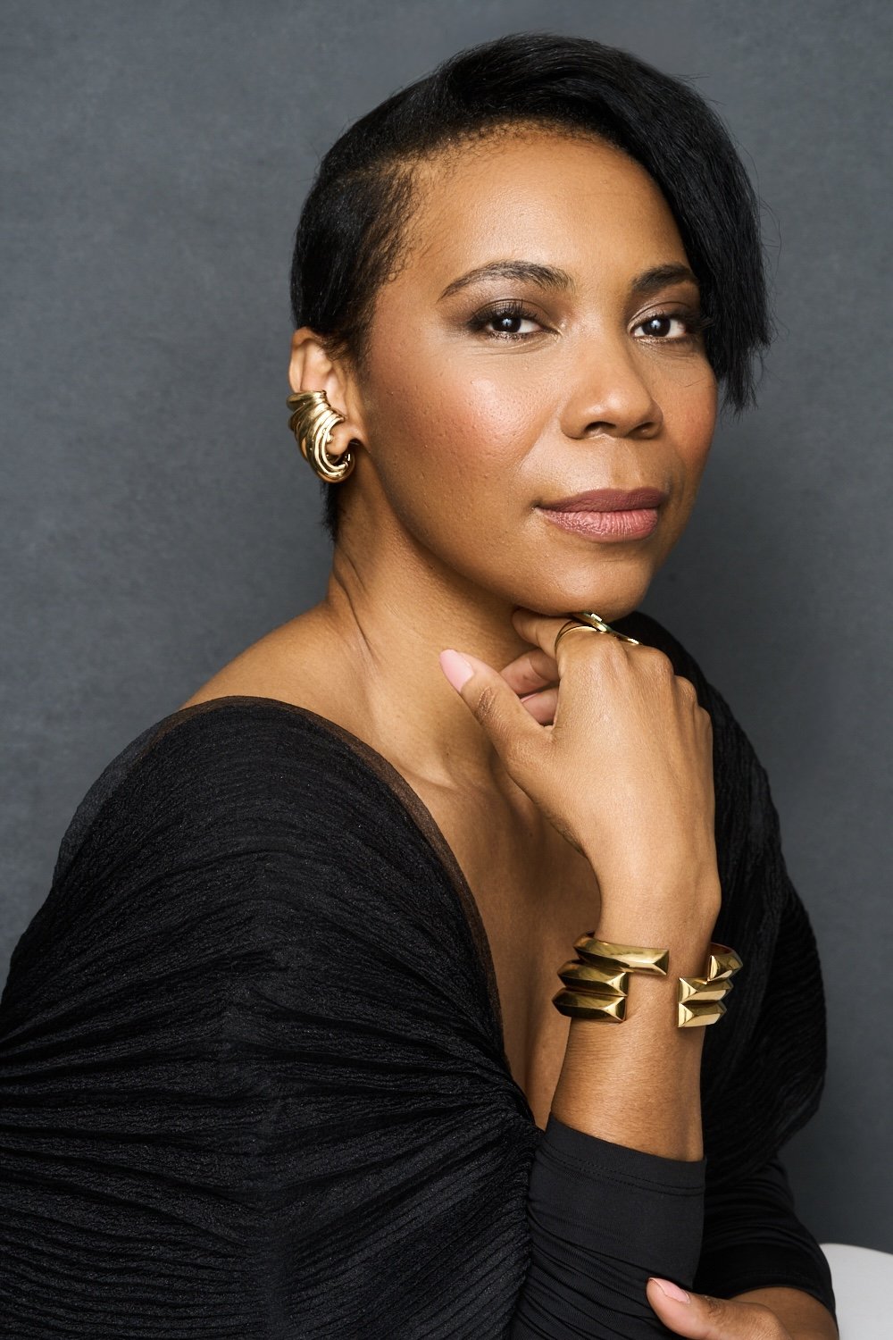 A woman with short black hair, wearing gold jewelry including earrings, bracelet, and ring, poses against a gray background, wearing a black top.