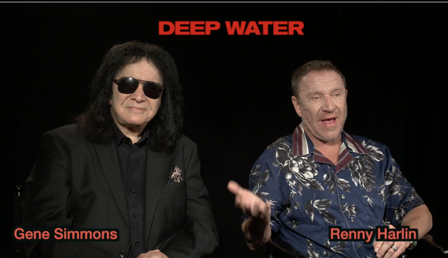 Gene Simmons and Renny Harlin sitting and talking during an interview or discussion, with the text "Deep Water" above them. Gene Simmons is wearing sunglasses and a black suit, while Renny Harlin is wearing a blue Hawaiian style shirt.