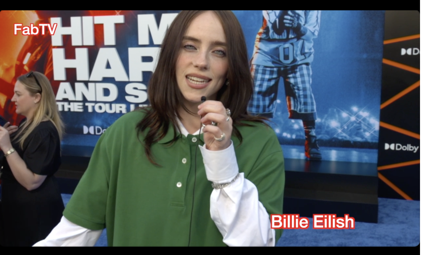 A woman with dark brown hair wearing a green polo shirt over a white long sleeve shirt, holding a microphone and smiling. In the background, another woman with blonde hair and glasses is seen in profile, also holding a microphone, with a sports-themed poster displaying the words "HIT ME HARD AND S" and an athlete in a blue and white uniform. The logo "FabTV" is in the top left corner, and the name "Billie Eilish" appears in red text at the bottom right.