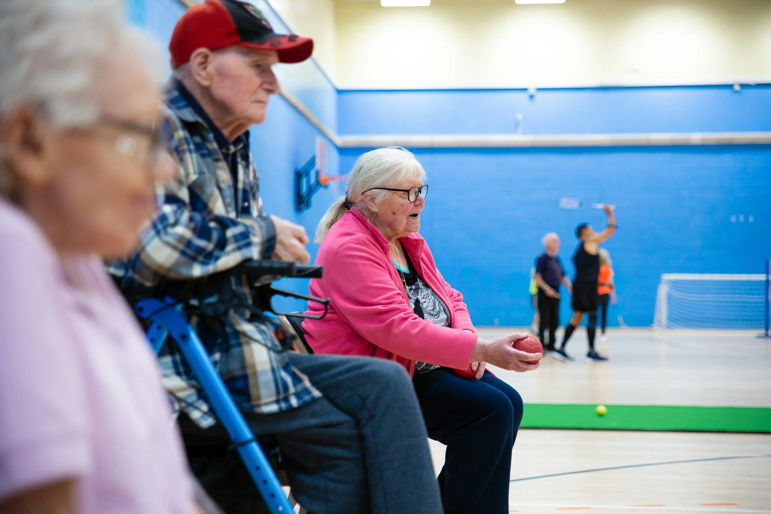 Photo showing older people sitting on a chair, using a seated frame to play bowls in a gym or leisure centre environment
