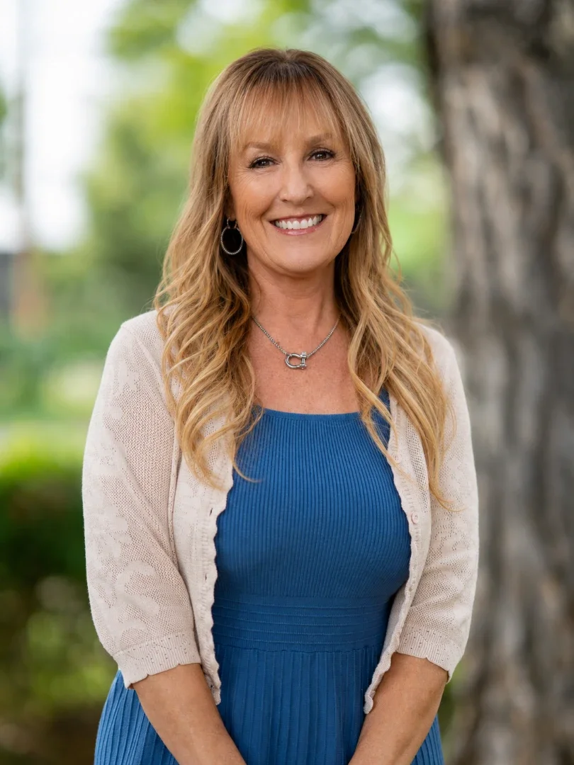 A woman with long, wavy blonde hair smiling outdoors, wearing a blue dress, a white cardigan, and jewelry, with trees and greenery in the background.
