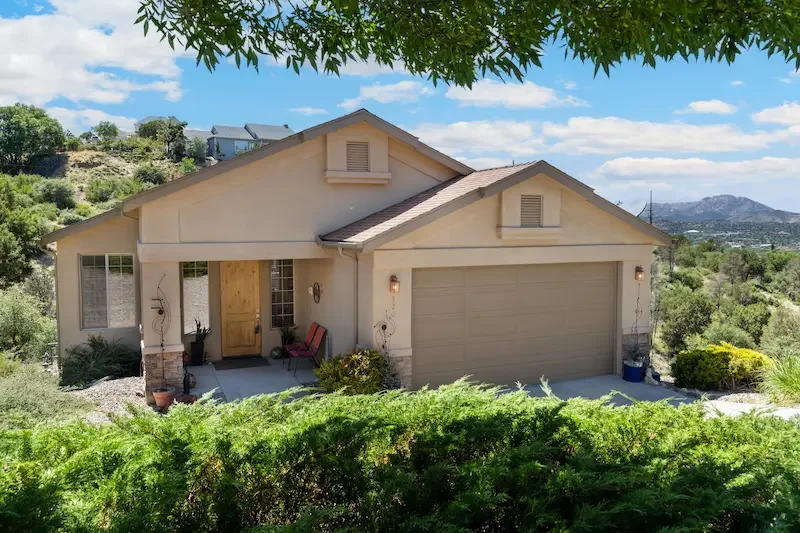Single-story house with attached garage, front porch with seating, surrounded by greenery and hills in background.