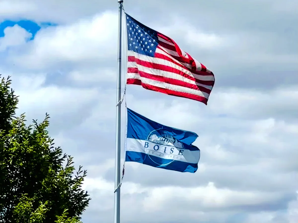 American flag, Boise city flag, and a tree with green leaves, all flying on a flagpole against a cloudy sky.