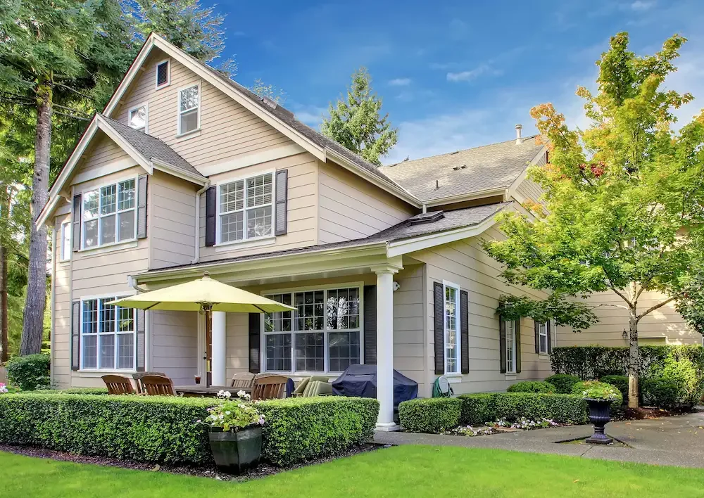 Large beige two-story house with multiple windows, surrounded by green trees and manicured bushes, with a back patio area that has outdoor chairs, a table with an umbrella, and a small garden, under a blue sky.