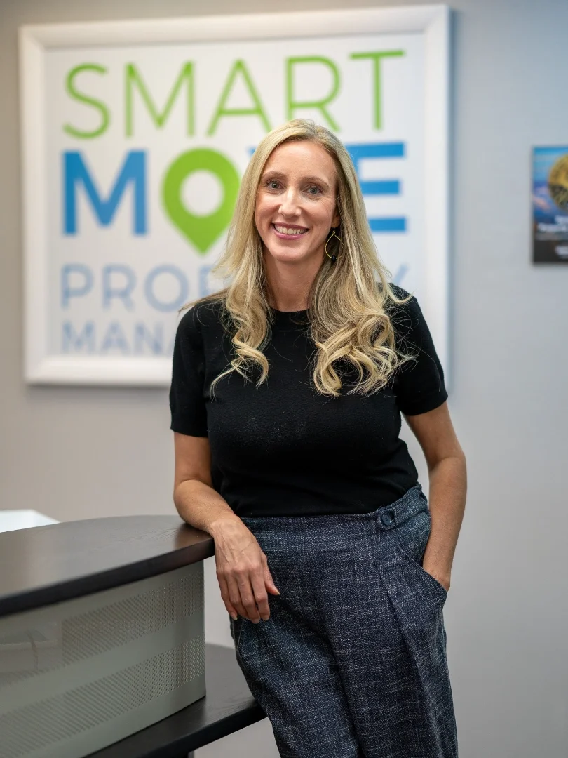 A woman with long blonde hair smiling, wearing a black top and gray checkered pants, standing in front of a wall with a colorful sign that says "Smart Move" and additional text.