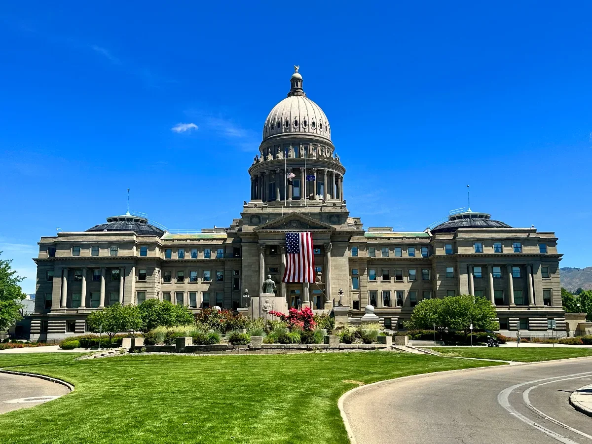 The image shows the Utah State Capitol building with a large American flag hanging in front. The building has a prominent dome and is surrounded by well-maintained green lawns, trees, and a clear blue sky.