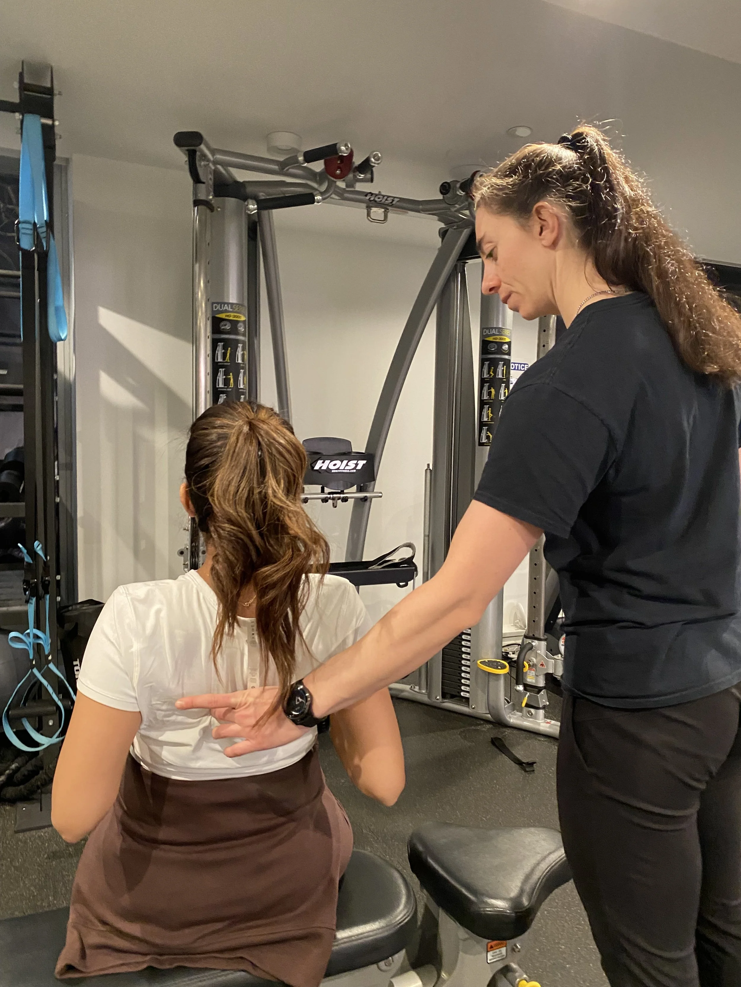 A woman with long brown hair in a black shirt is standing next to a seated woman with long hair and a white shirt, in a gym or fitness center, with exercise equipment and straps in the background.