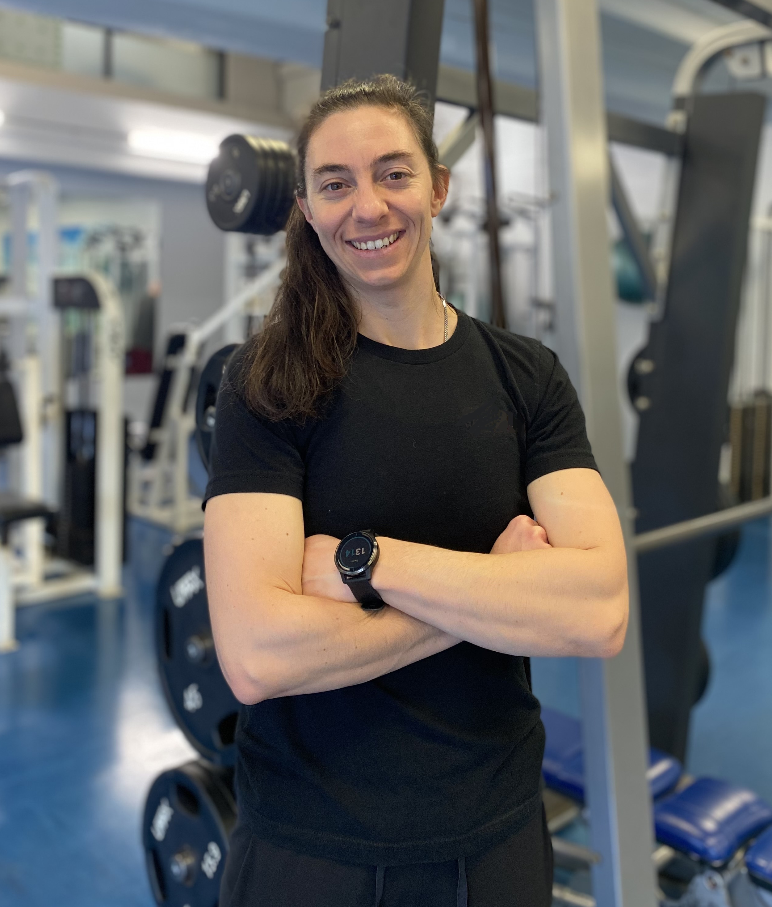 Smiling woman with brown hair in a ponytail, wearing a black T-shirt, standing in a gym with weight machines in the background.