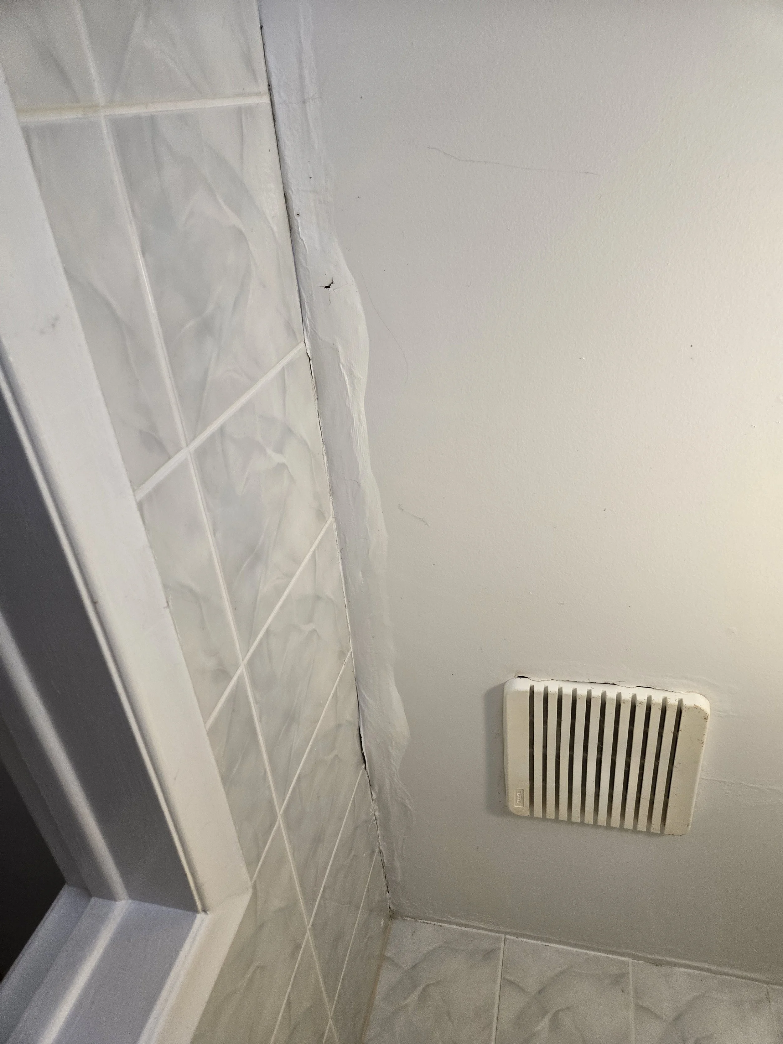 Close-up view of a tiled floor and wall in a bathroom or kitchen, with a white vent cover on the wall and some exposed caulk or sealant along the edge of the tiles.