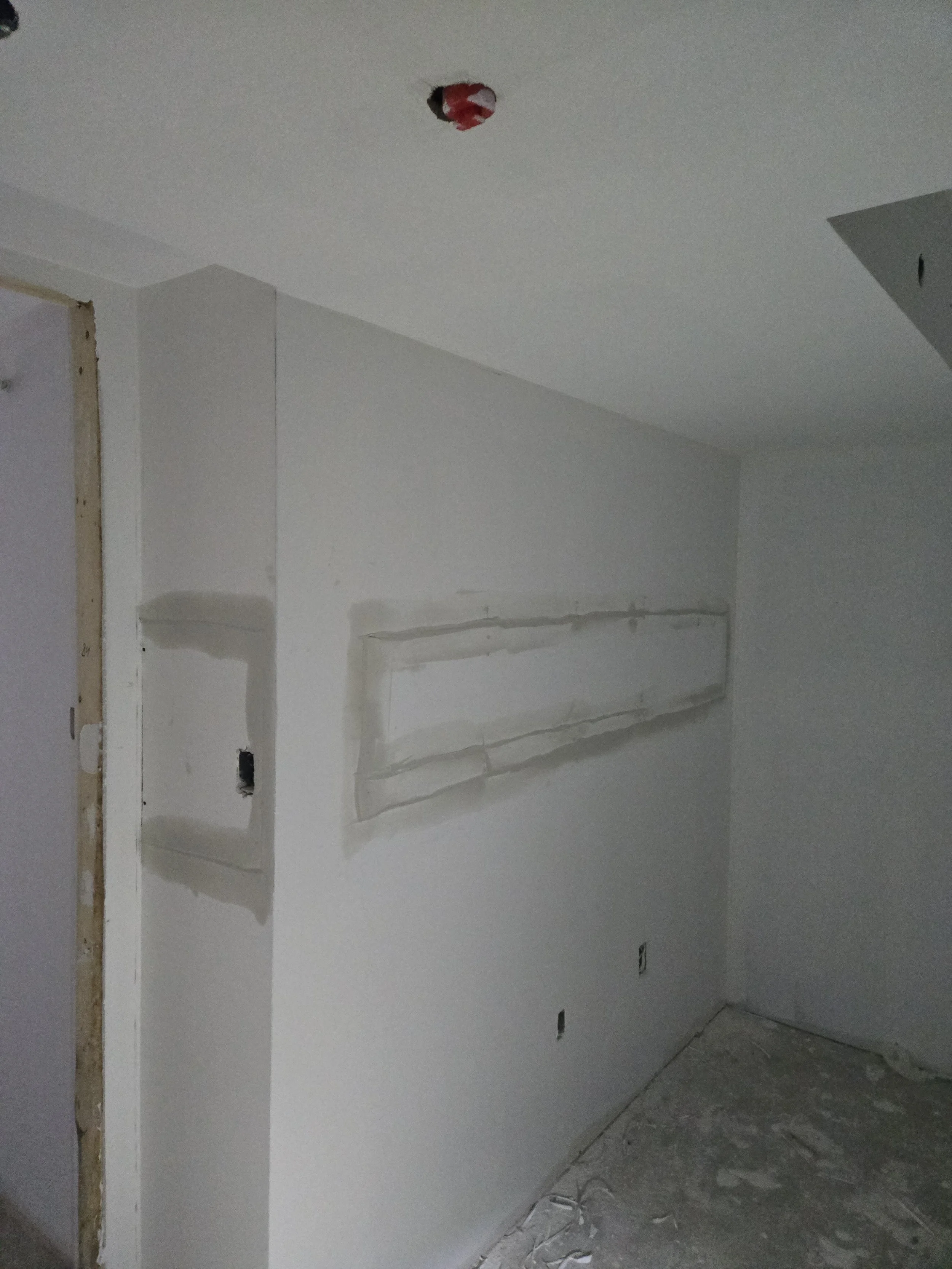 Room under construction with white painted walls, a rectangular hole in the wall covered with plastic, and construction debris on the floor.