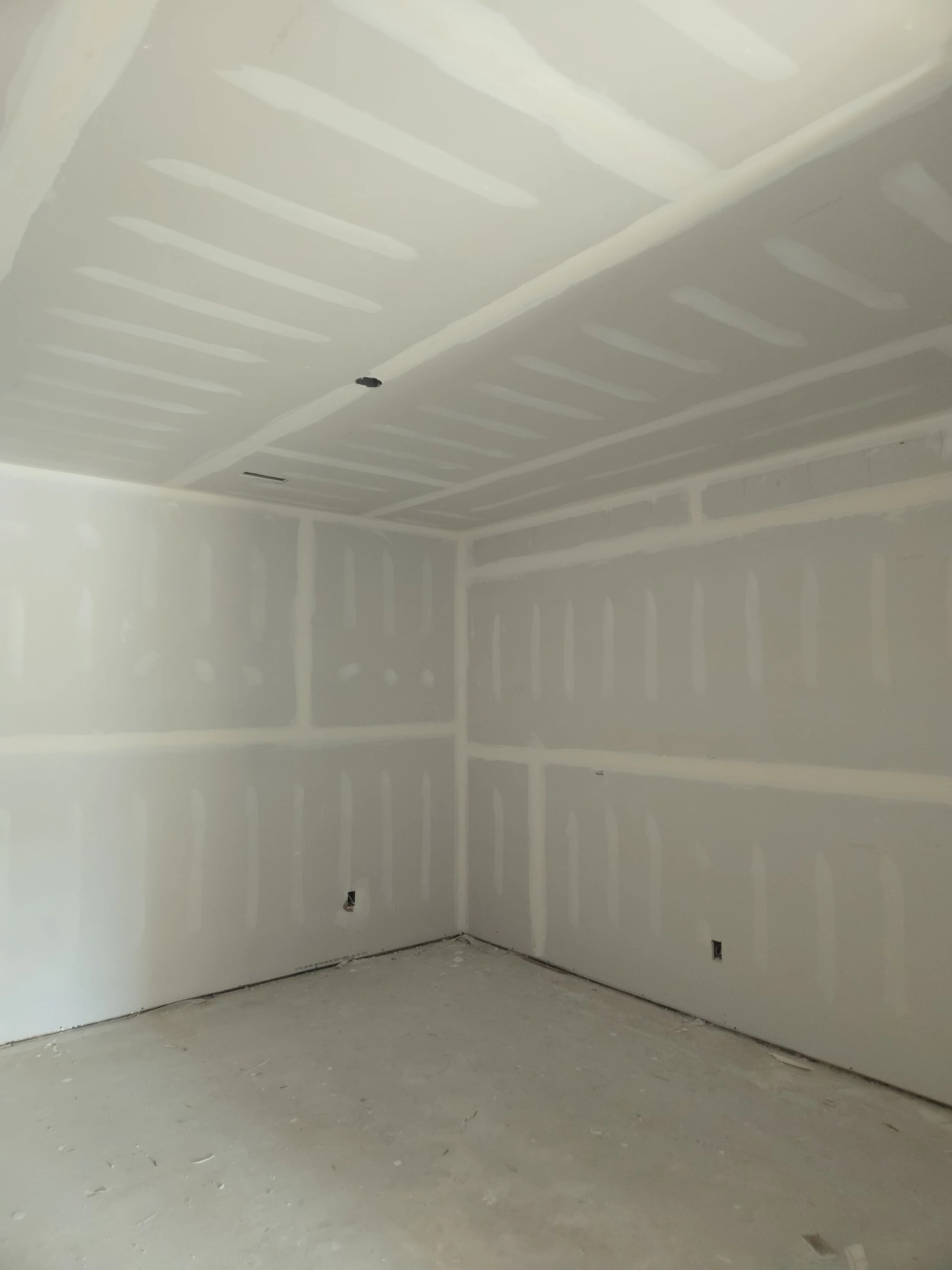 Empty room under construction with drywall walls and ceiling, with visible joints and some electrical outlets.