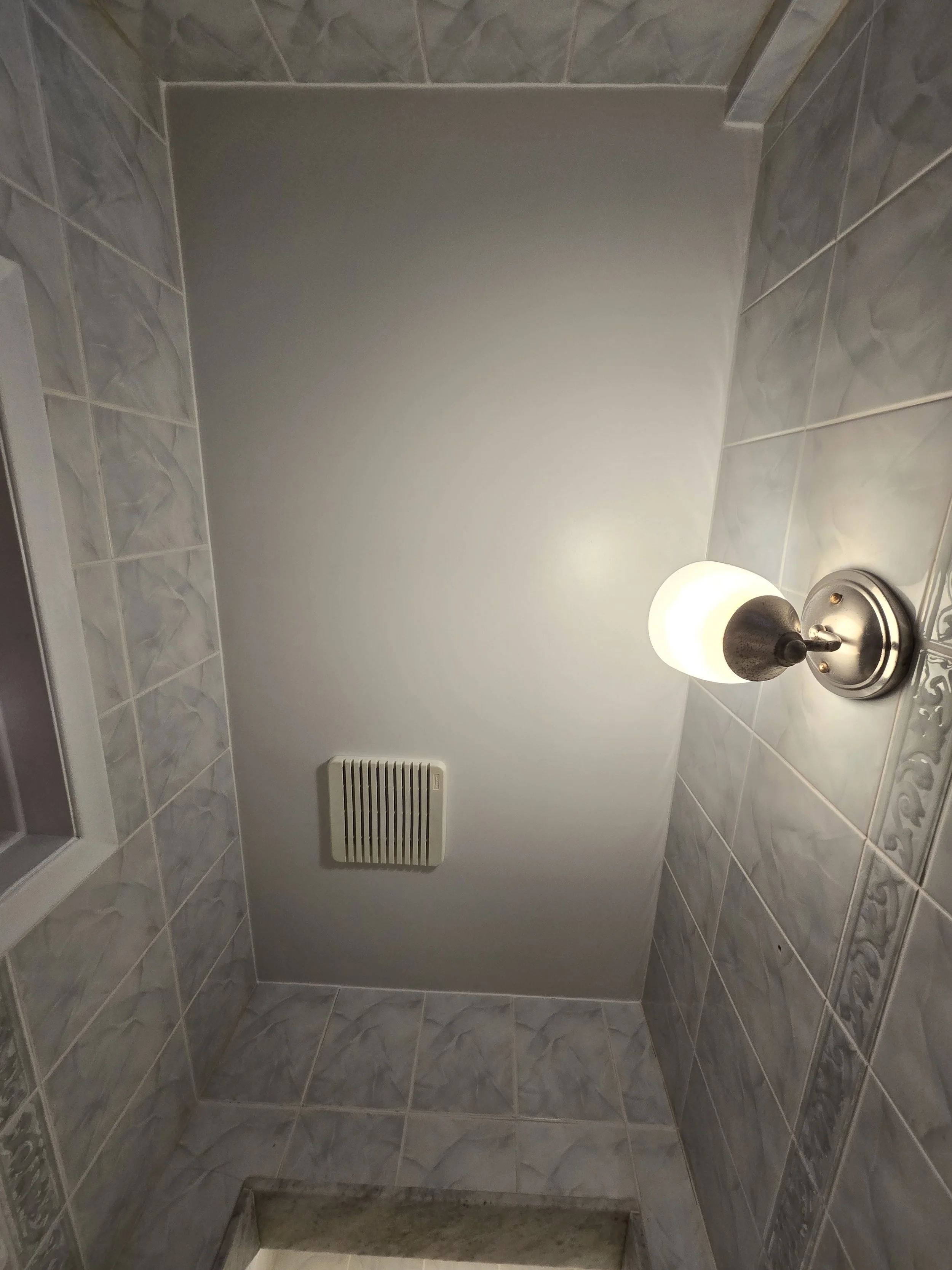 A small bathroom with gray tiled walls, a white ceiling vent, and a white light fixture with a frosted glass shade.