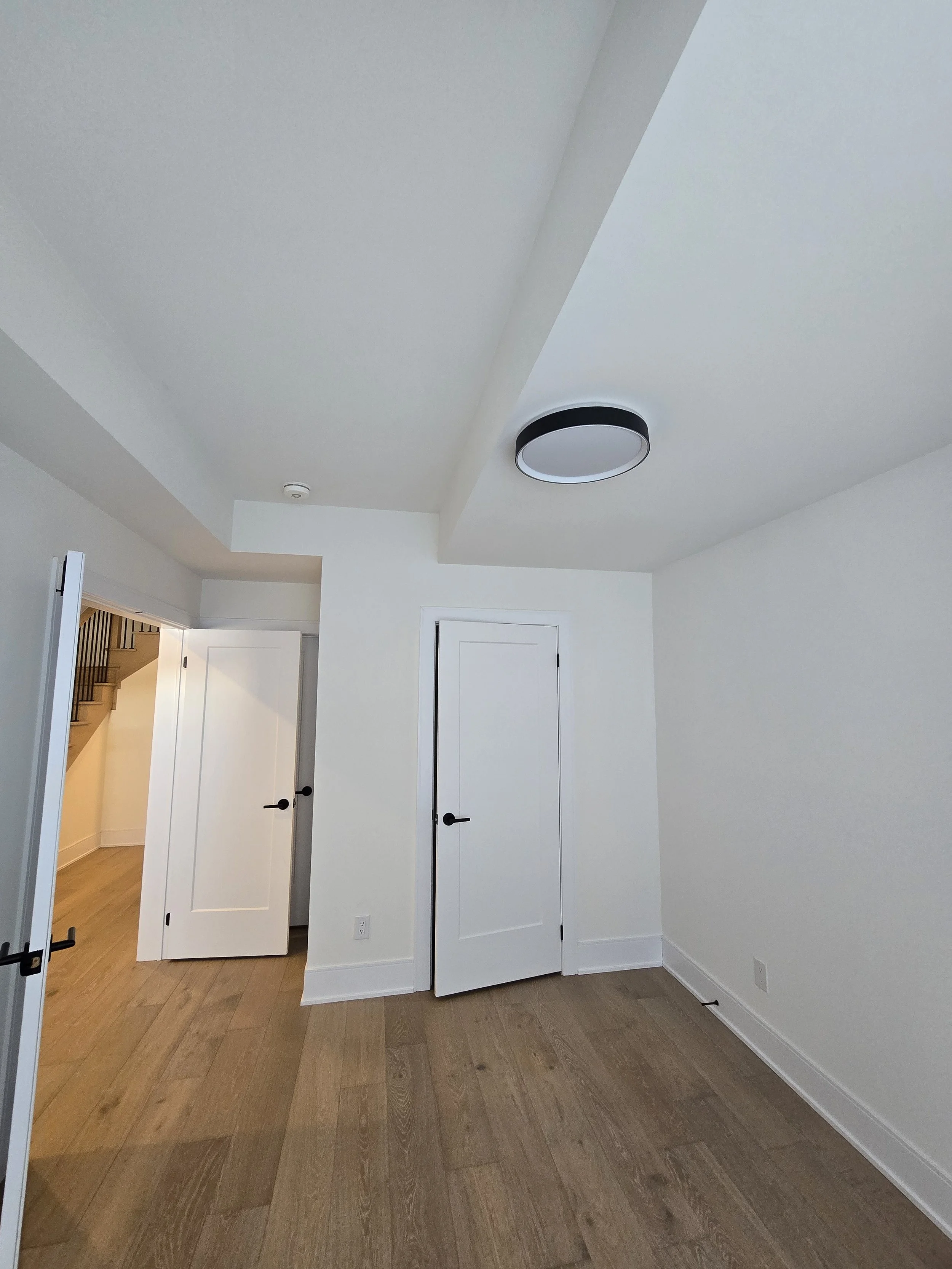 Empty room with white walls, wooden flooring, a ceiling light fixture, and open doors leading to a hallway with a staircase.