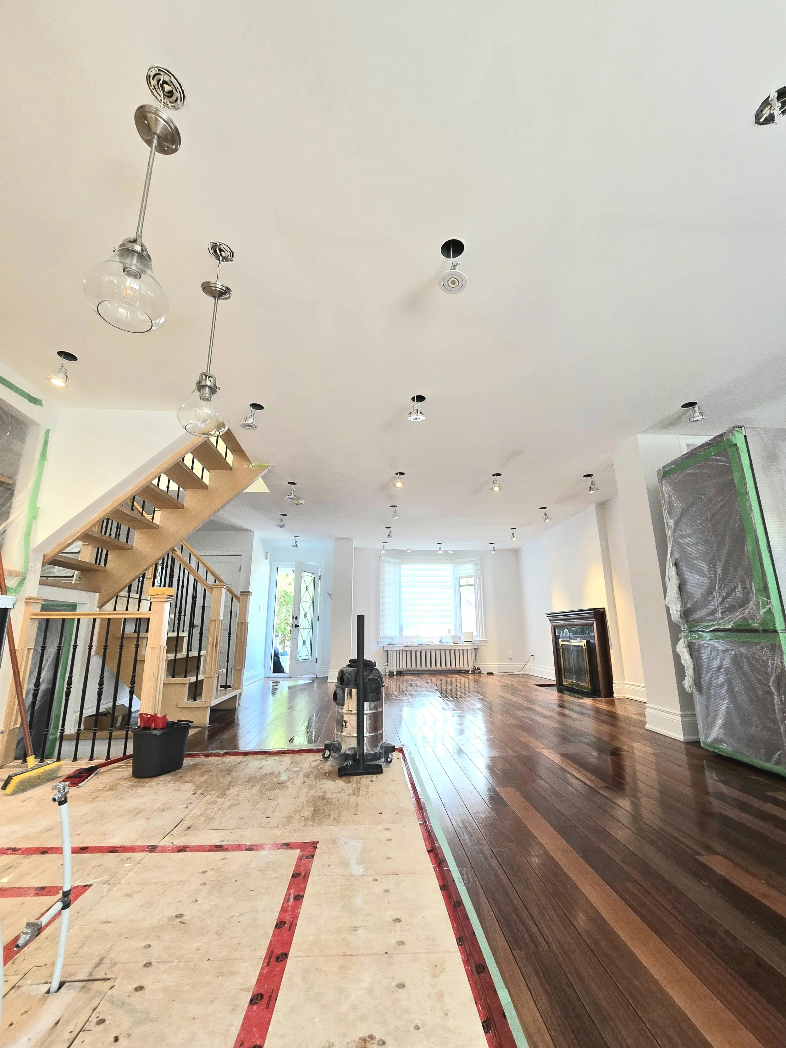 Living room undergoing renovation with hardwood flooring, partially removed flooring area, staircase with wooden steps and black metal balustrade, multiple ceiling lights, large window, green painter's tape along edges, stairs, and construction equip