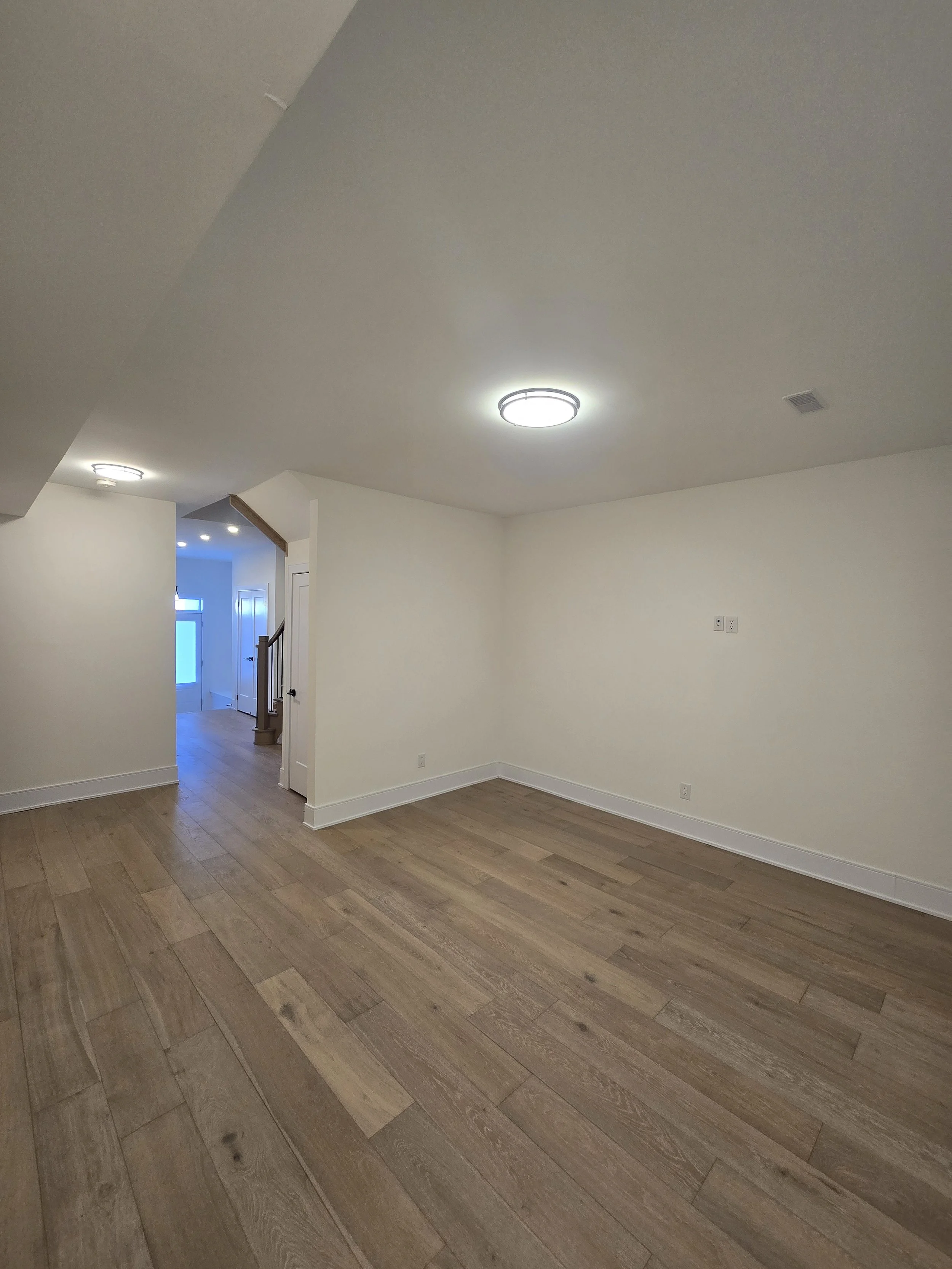 Empty living room with wooden floors, white walls, ceiling lights, and a hallway with stairs leading to the entrance door.