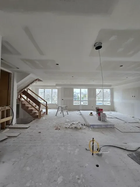 Interior of a room under construction with unfinished walls, ceiling, and flooring, along with construction tools and materials. A wooden staircase is on the left, and large windows are at the back.
