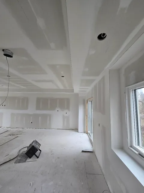 Interior of a room under construction with drywall installation, two large windows, and a partial ceiling with cutouts for lighting.