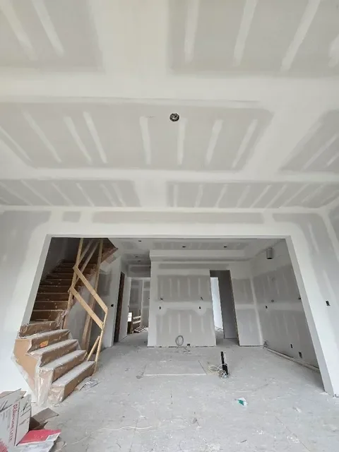 Interior of a house under construction with drywall walls and ceiling, staircase on the left side, and construction materials on the floor.