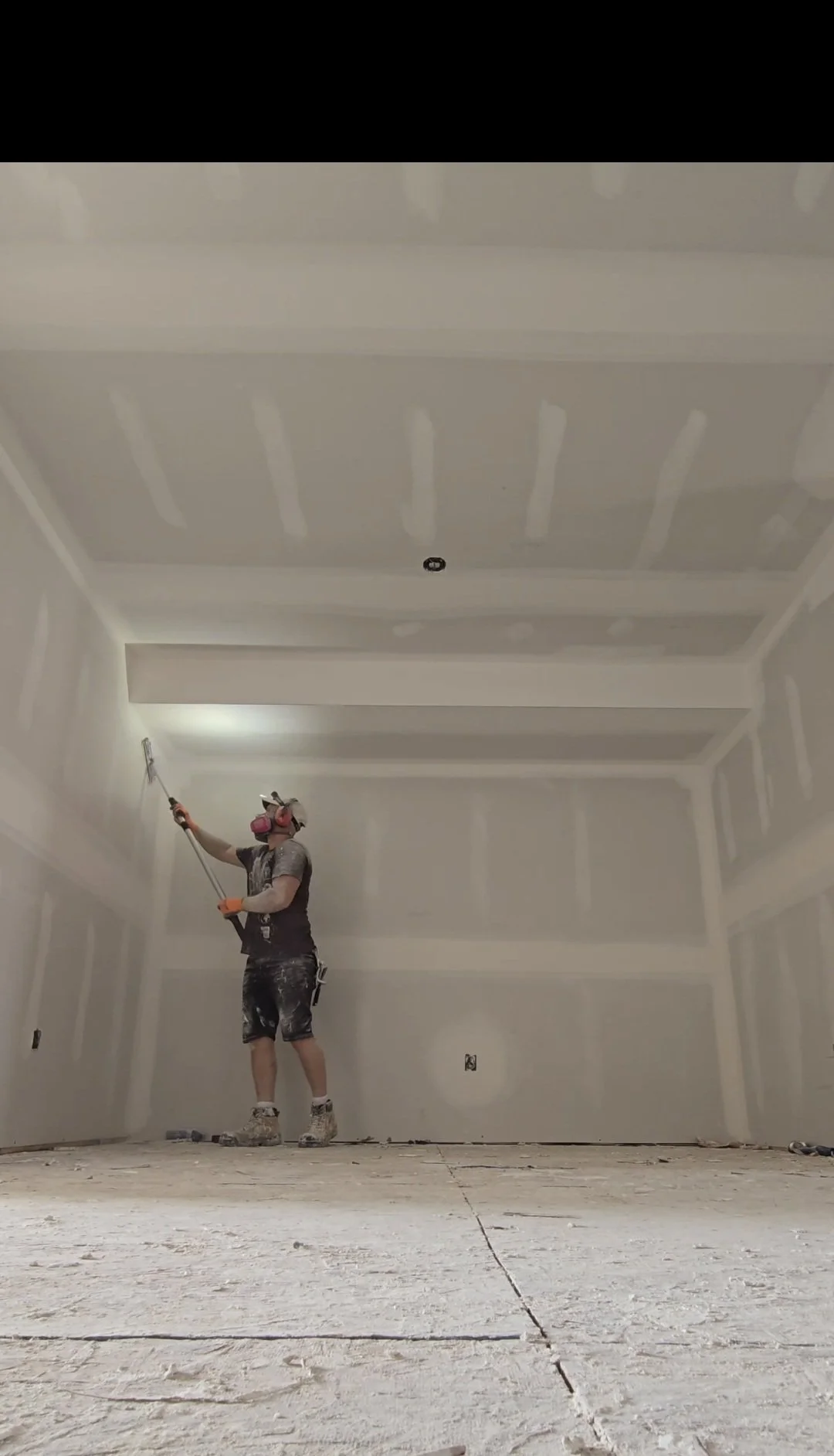 A person wearing a protective mask and gloves is standing on a ladder inside a room, spraying or painting the walls.