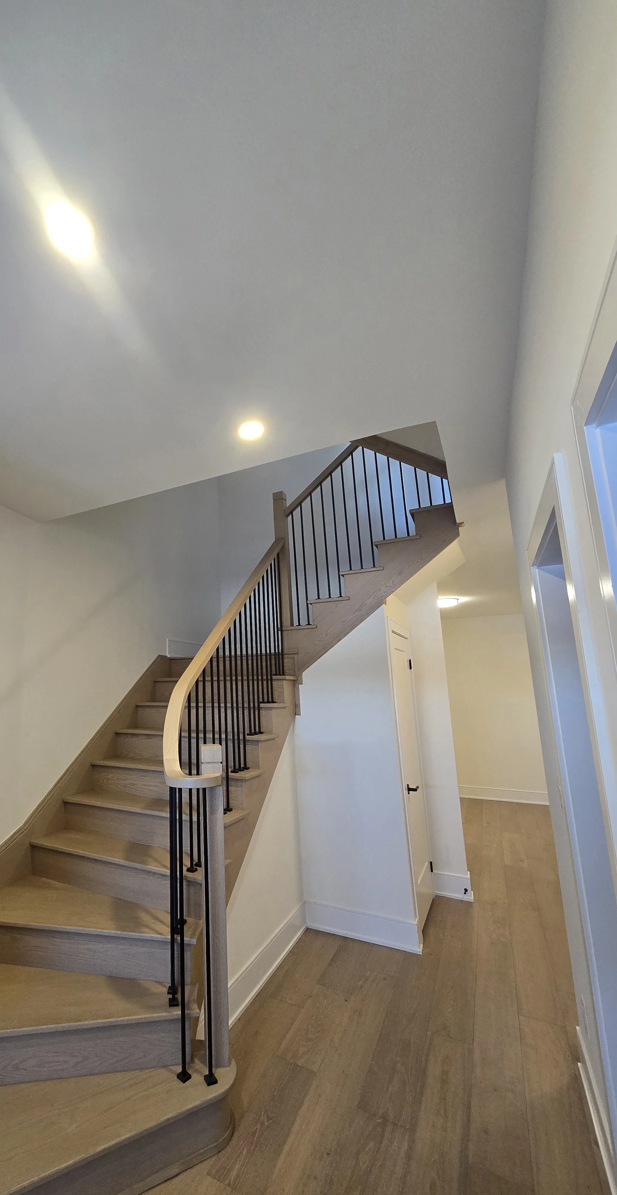 Interior view of a wooden staircase with black railings leading to the upper floor, with white walls and a wooden floor, under ceiling with recessed lights.