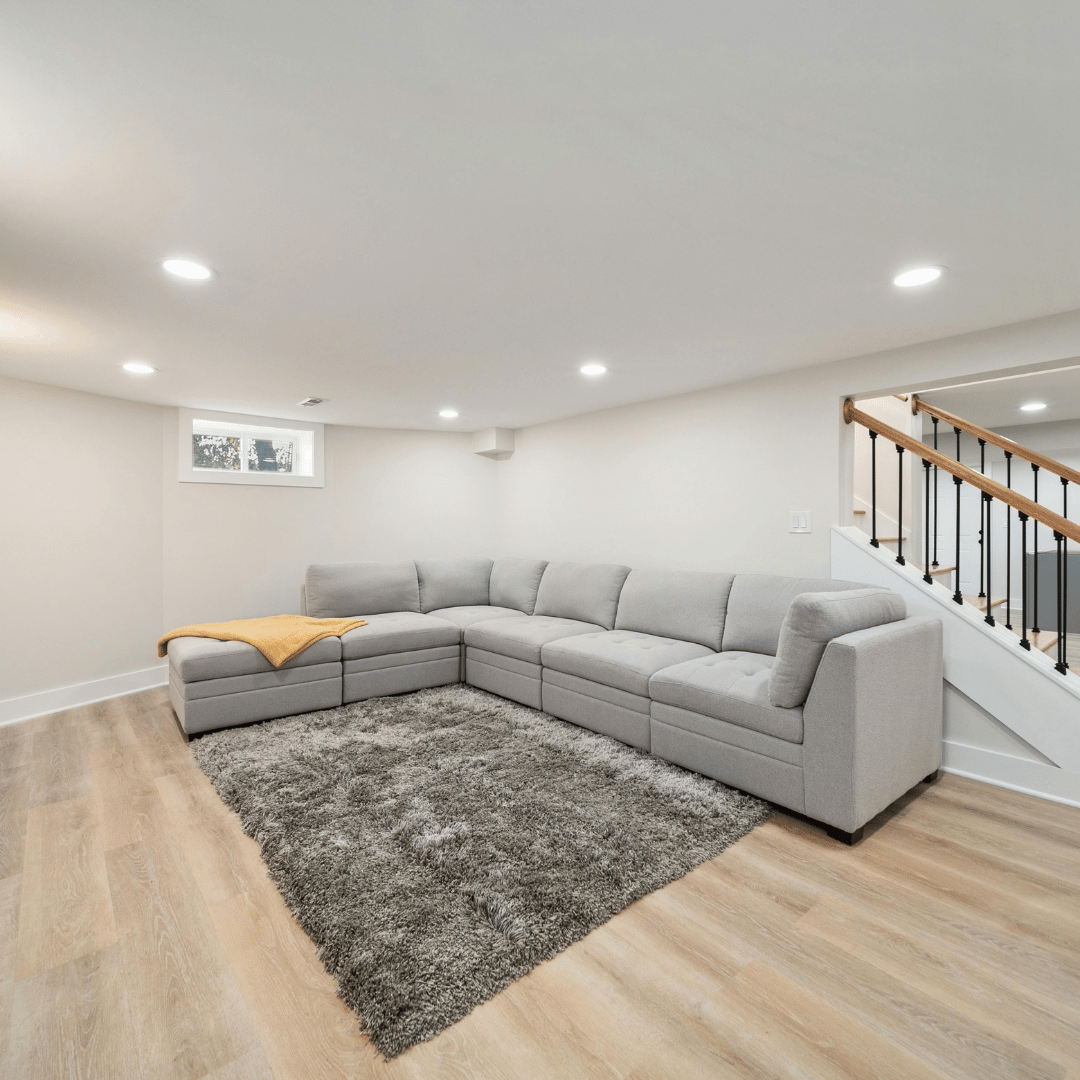 A basement living room with a large gray sectional sofa, a plush gray rug, light wood floor, white walls, small window, and a staircase with black balusters and wooden handrail.