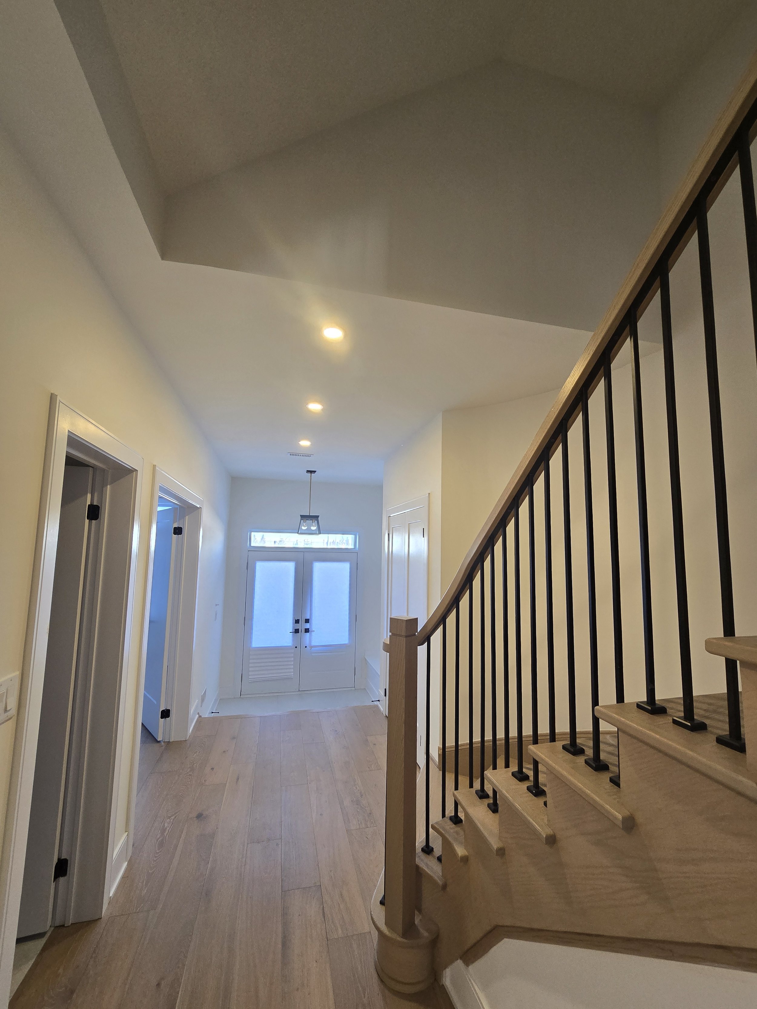 Interior view of a hallway with light-colored walls, hardwood flooring, a front door with a window, multiple doorways, and a staircase with a wooden handrail and black balusters.