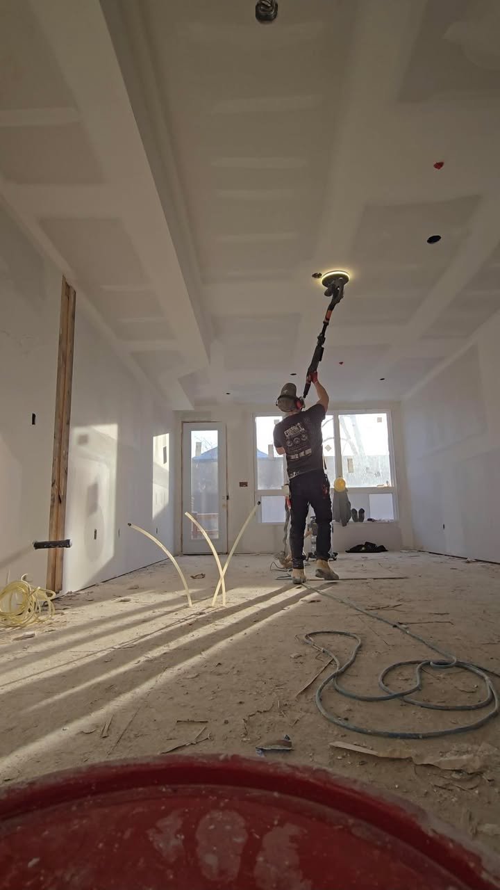 Construction worker in a cap and t-shirt using a tool to install drywall in a room with large windows, unfinished floor, and overhead ceiling lighting fixtures.