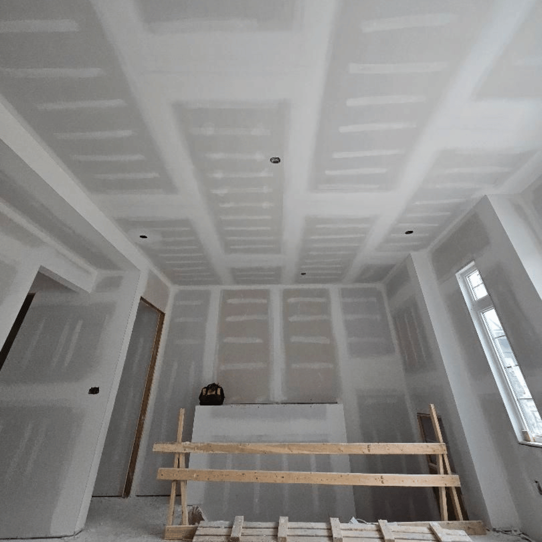 Interior of a room under construction with drywall installed, unfinished walls and ceiling, and construction materials on the floor.