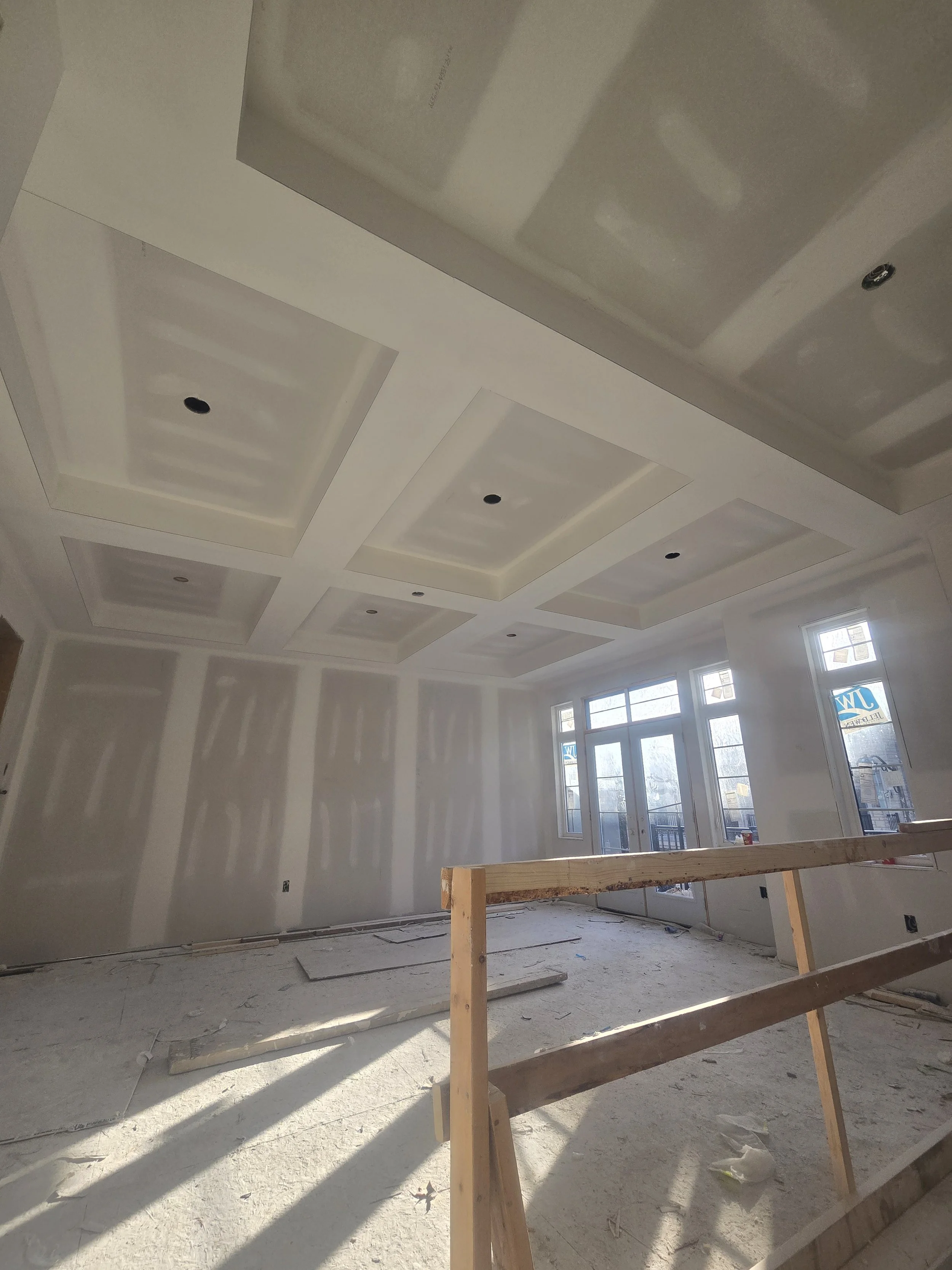 Interior of a house under construction with a tray ceiling, drywall walls, unfinished floor, and large windows