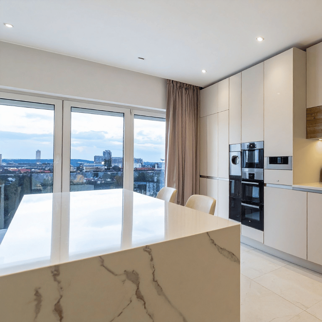 Modern kitchen with a marble island, built-in ovens, large windows showing cityscape, beige cabinets, and beige chairs.